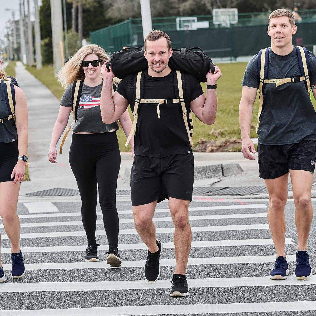 Group of people rucking with GORUCK backpacks crossing a street, wearing athletic gear outdoors