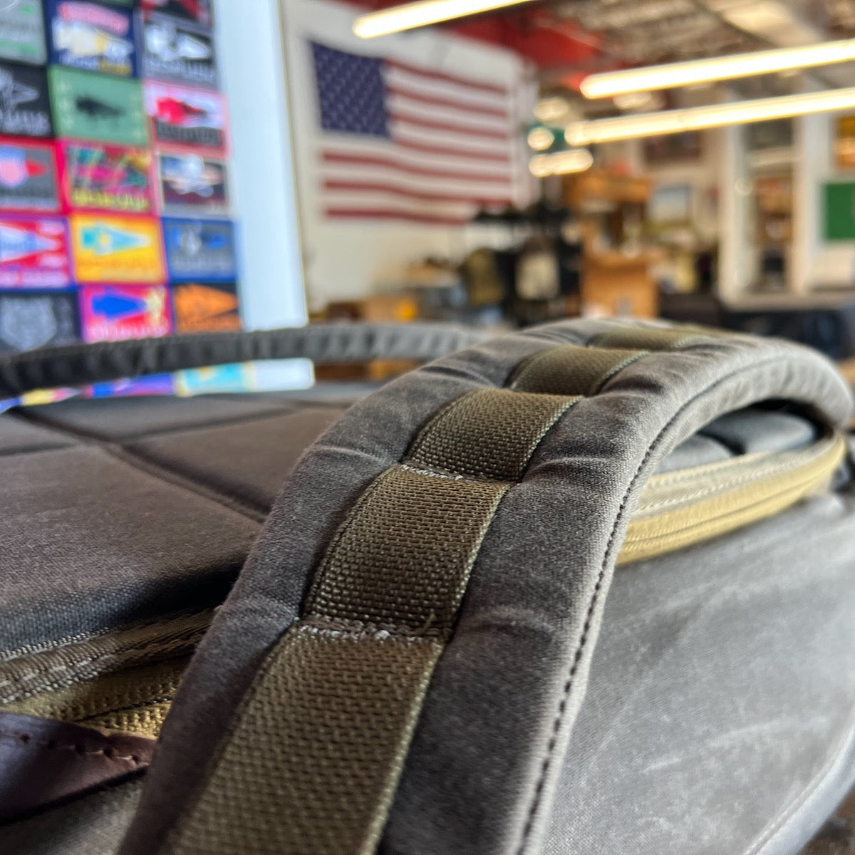 Close-up of a padded backpack strap from the GR1 USA - Heritage Waxed Canvas rucksack, with an American flag and colorful signs blurred in the background.
