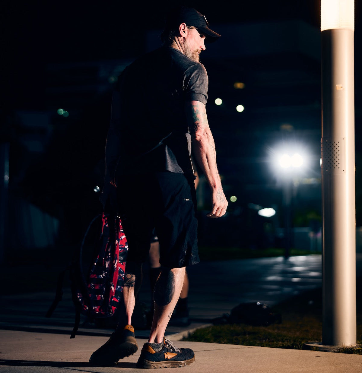 A bearded man with tattoos wears a cap, t-shirt, and technical cargo shorts while walking on a sidewalk at night, carrying a red patterned backpack.