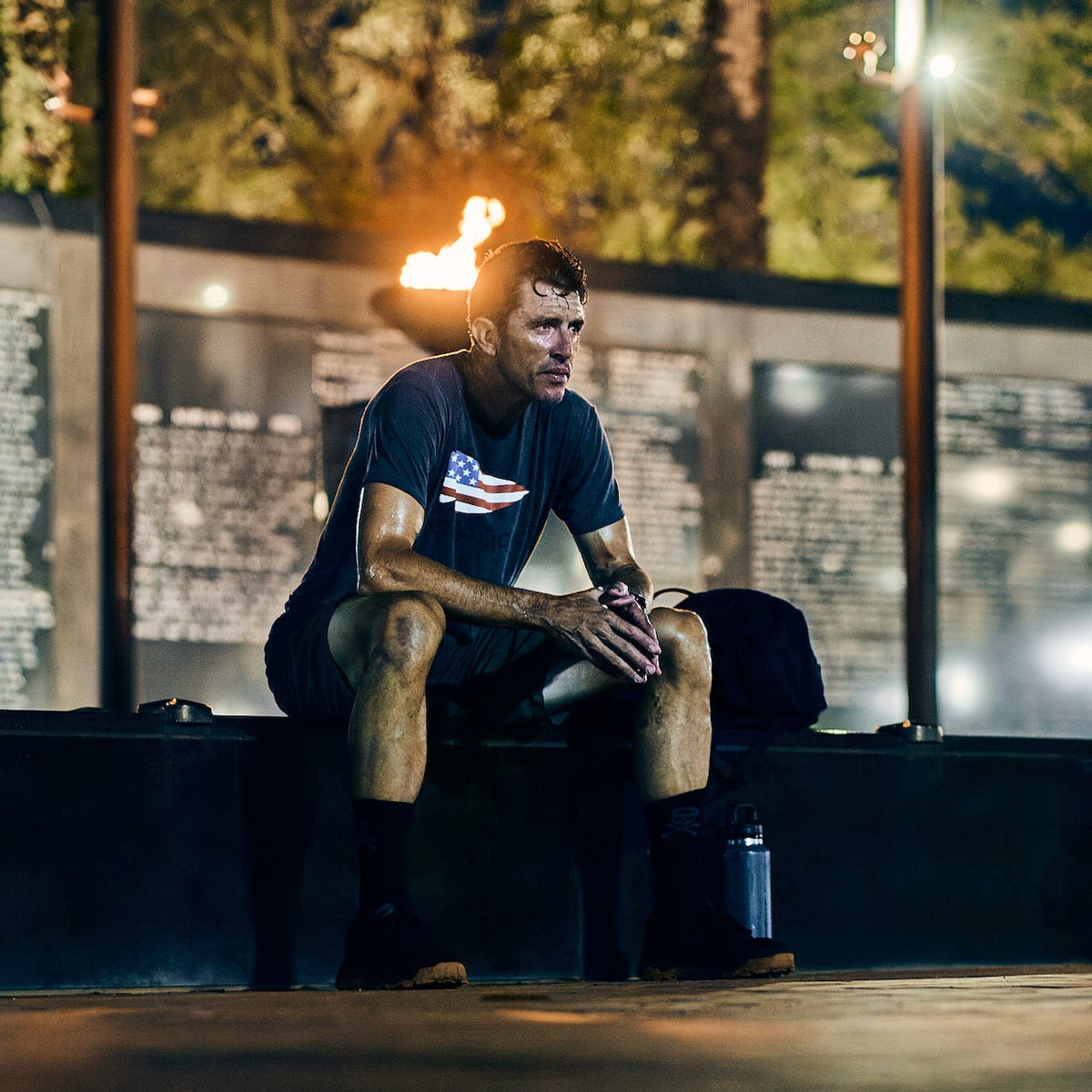 A sweaty man in an American flag t-shirt and tough shorts sits on a ledge in front of the Vietnam Veterans Memorial during warm weather rucking.