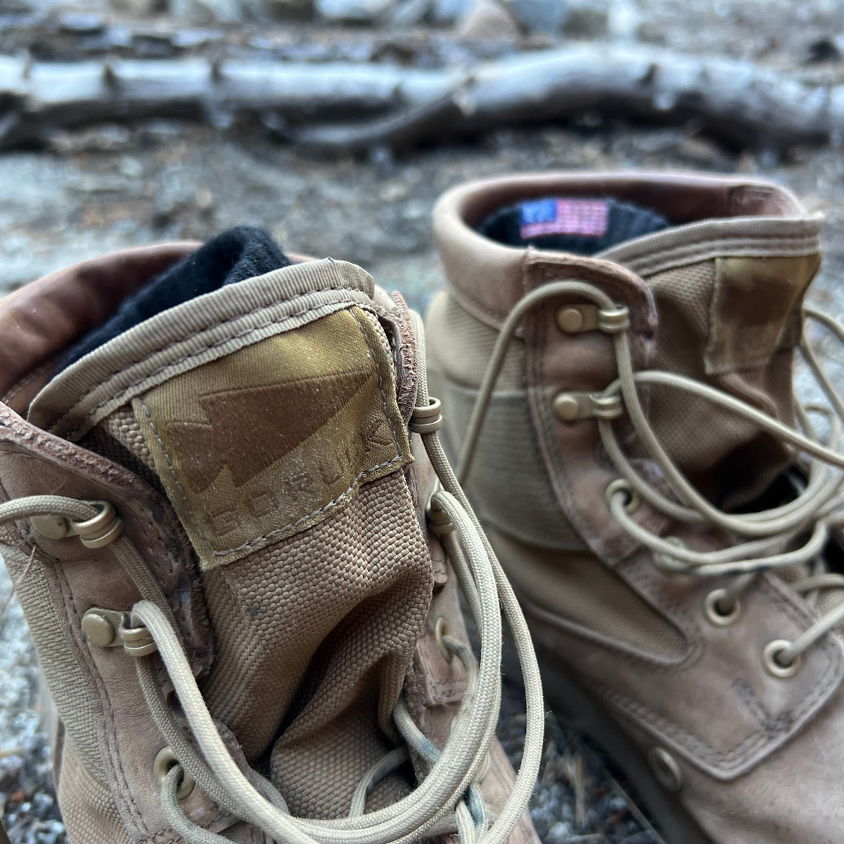 A close-up of tan MACV-2 Mid Top boots in Briar + Coyote with untied laces on a rocky outdoor surface.