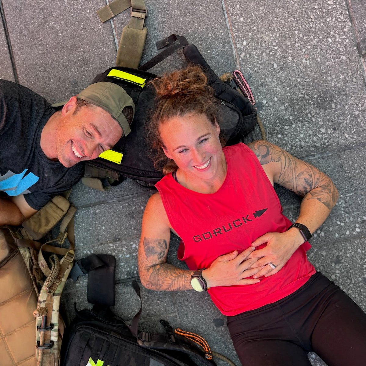 Two people smiling, lying on the ground with backpacks—one wearing the Women’s Performance Tank made from featherweight ToughMesh fabric in red, crafted in the USA by GORUCK, and the other sporting a cap.