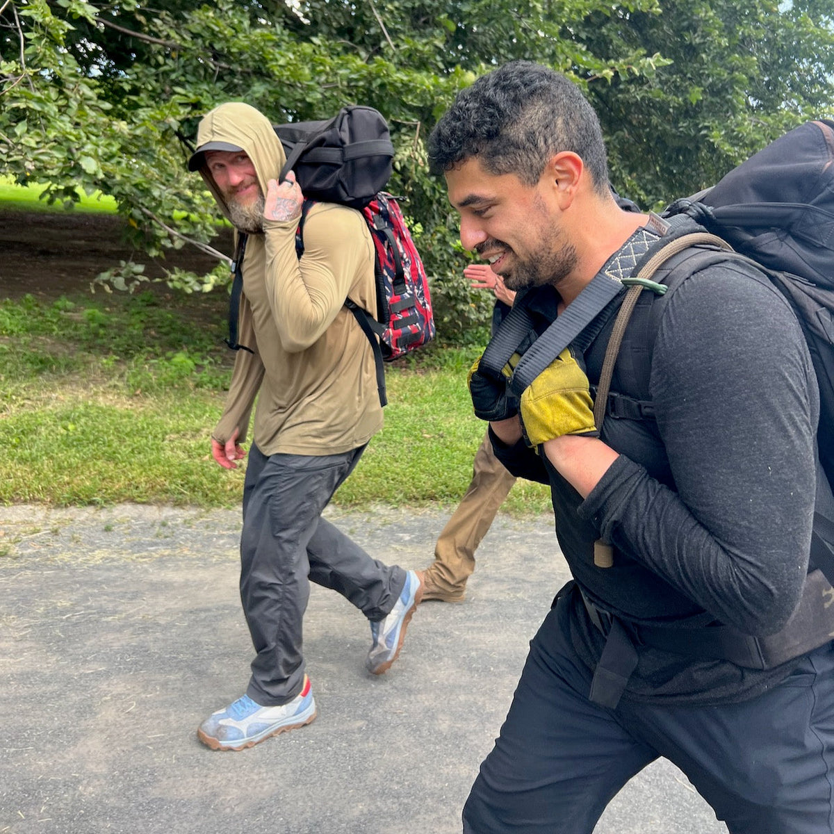 Two men with backpacks walk outdoors, smiling, on a paved path near grass and trees, showcasing the rugged durability of the Mackall 45 – Americana and its Special Forces rucking chassis.