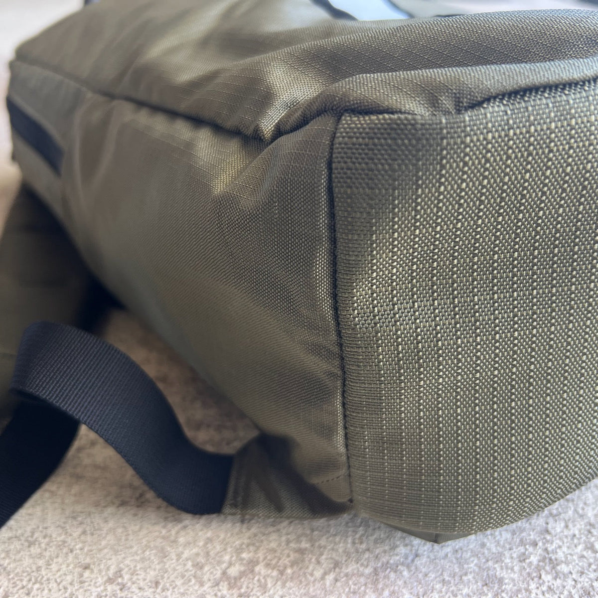 Close-up of the bottom corner of the Basic Rucker backpack in green with a black strap, resting on a light surface.