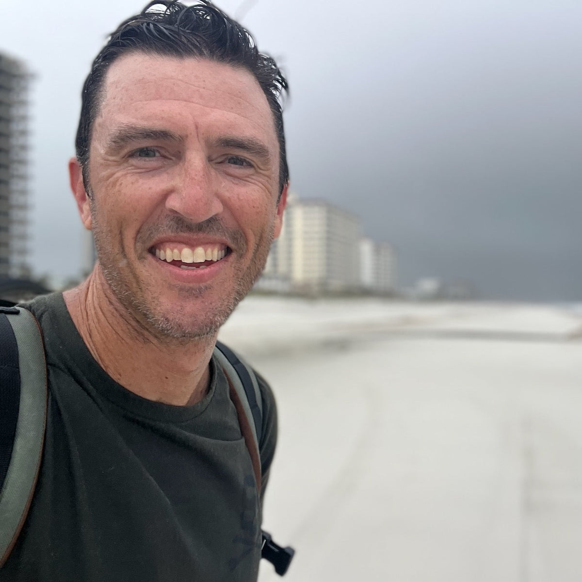 A smiling man with a GORUCK backpack stands on a beach with tall buildings in the background.