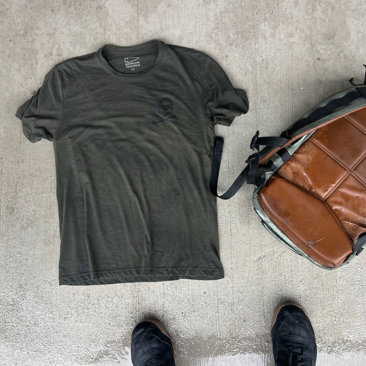 An olive green t-shirt with a skull and crossbones logo, a brown and camouflage backpack, and black shoes on a concrete surface, representing the GORUCK community.