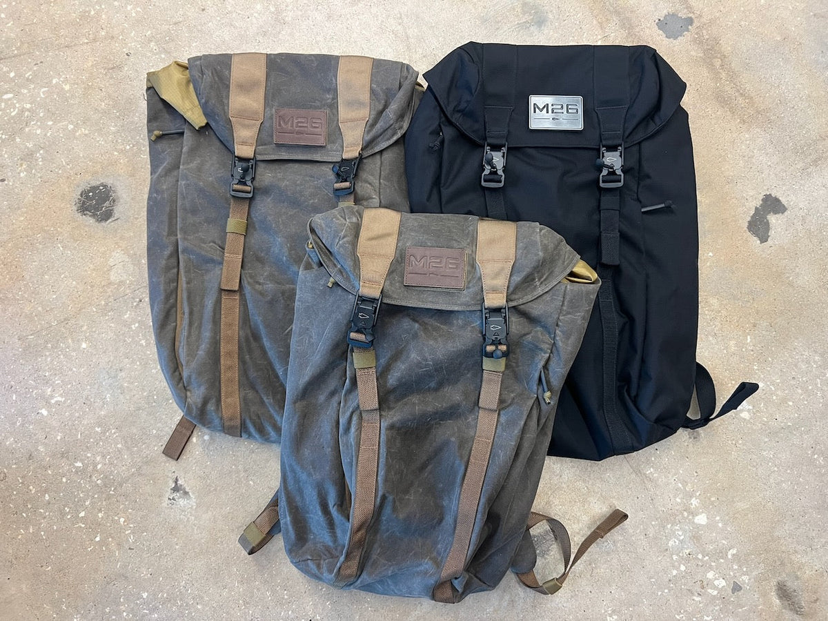 Three M26 field packs, including one solid black bag and two charcoal grey bags with tan straps, all featuring a quick access pocket.