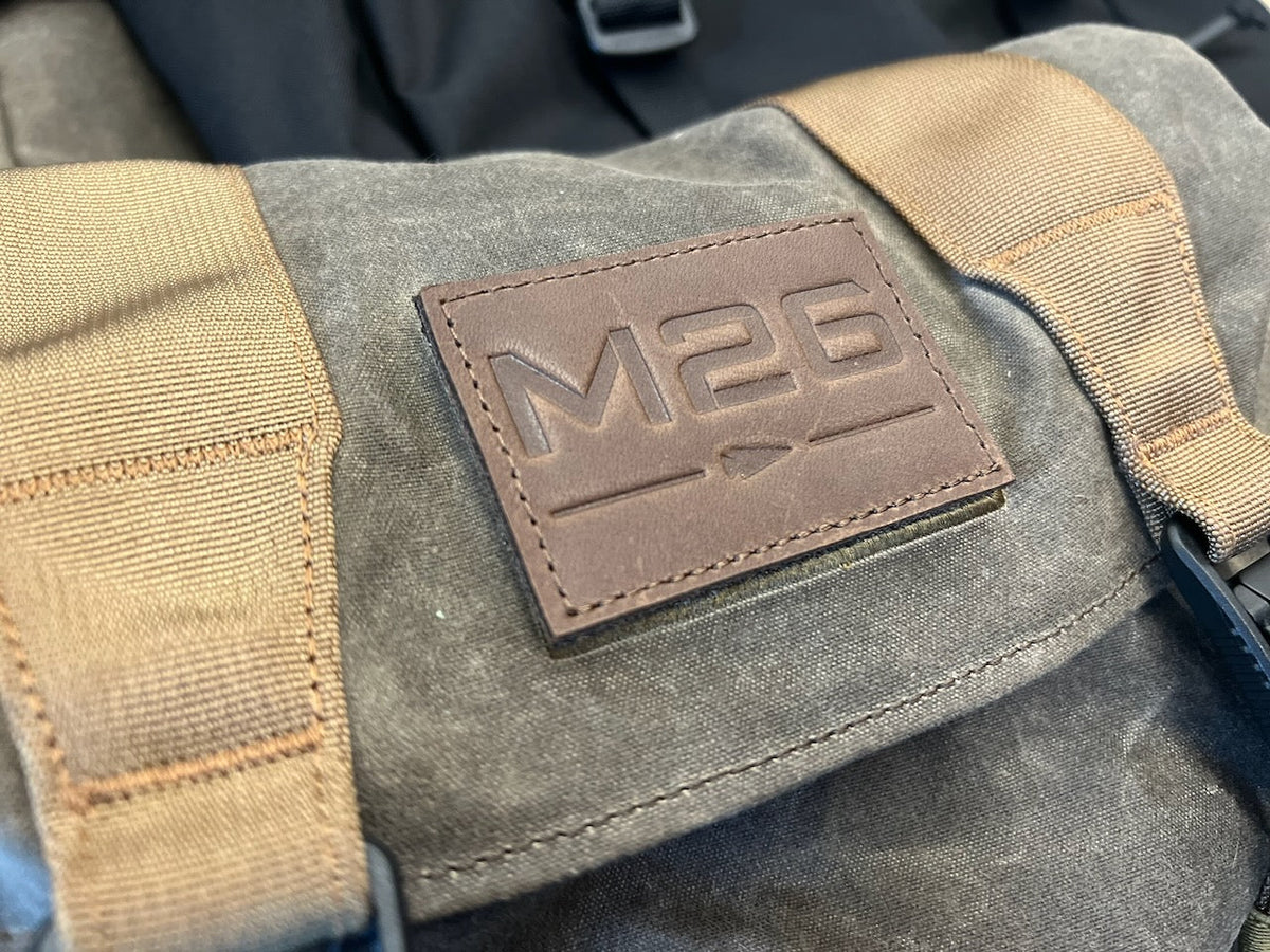 A grey WWII-inspired backpack with a brown leather patch embossed with the text “M26” and a horizontal arrow.