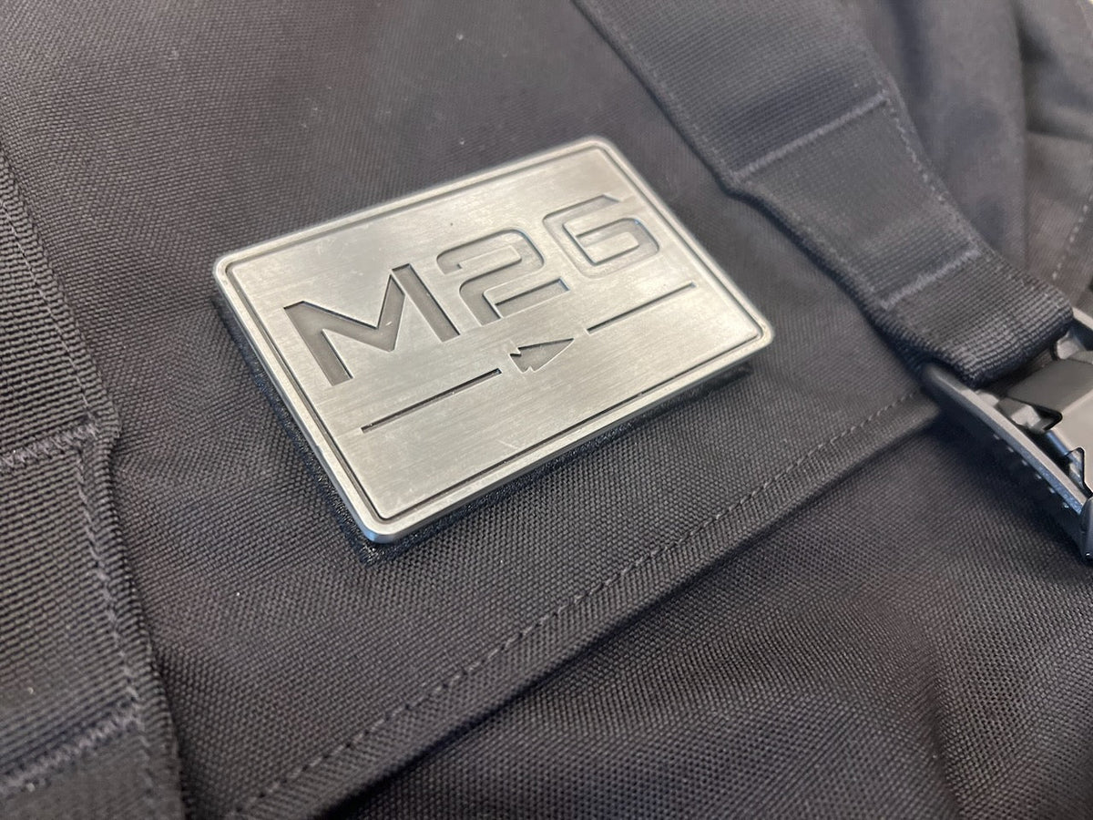 A black WWII-inspired backpack featuring a rectangular metal plate embossed with the text “M26” above a horizontal line with an arrow.