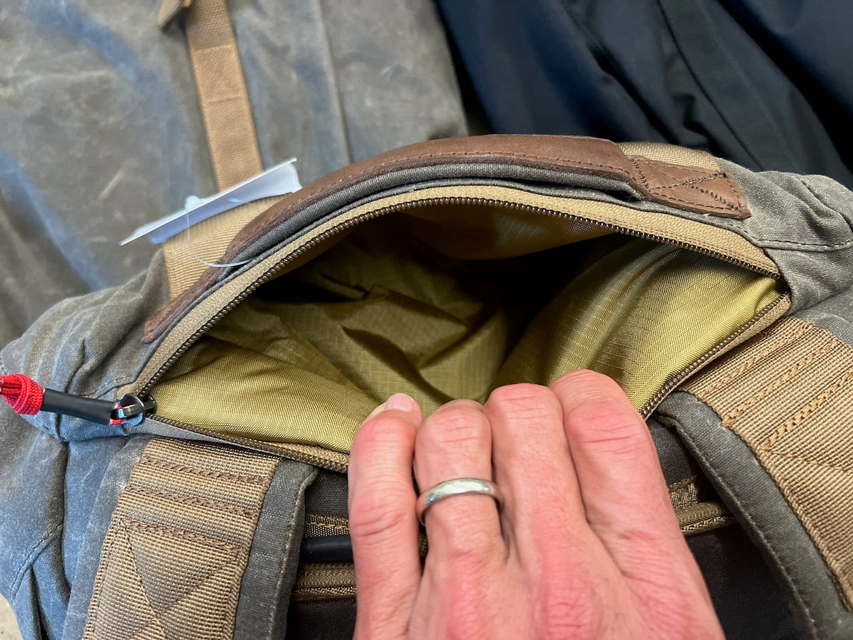 A hand pulls open the zippered quick access pocket of a grey canvas backpack, revealing a gold interior lining.