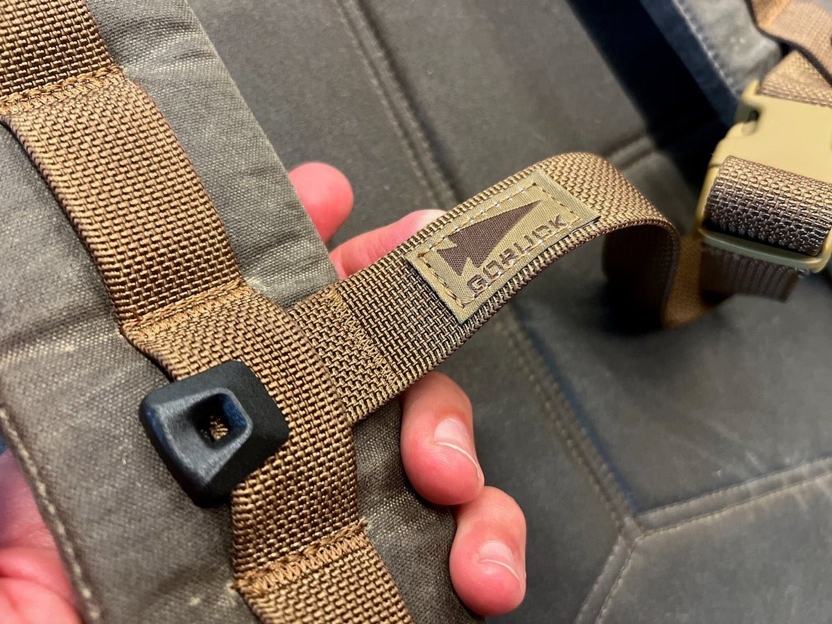 A person holds the brown webbing strap of a WWII-inspired GORUCK backpack, featuring a tan logo patch.