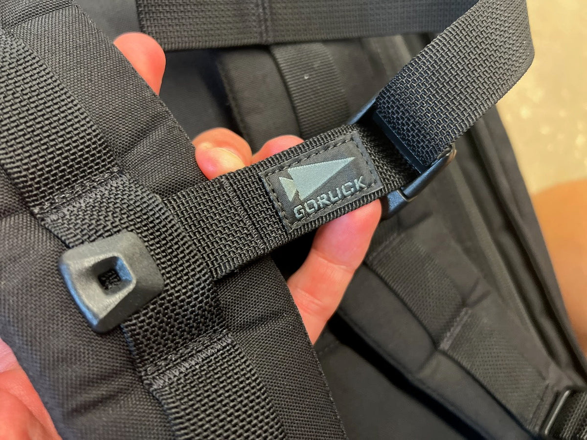 Close-up of a black GORUCK backpack shoulder strap featuring a small spearhead logo patch, styled after the M26 field pack.