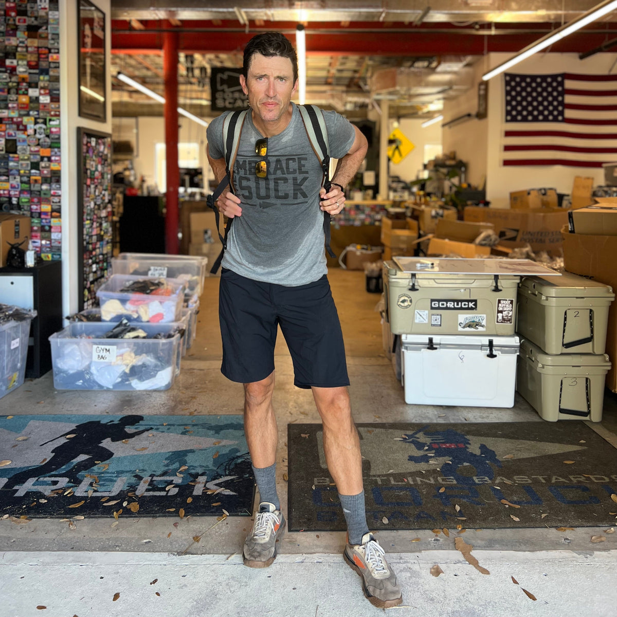 A man in athletic gear with a backpack stands in a warehouse of boxes and an American flag—ready for anything in Men’s Simple Shorts - Lightweight ToughDry®, featuring fast-drying, 2-way stretch fabric.