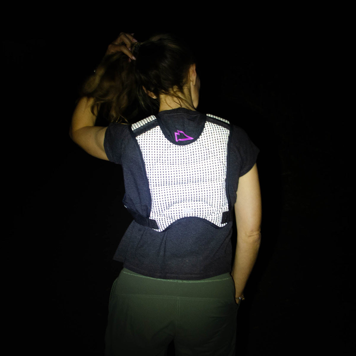 At night, a woman wearing the Spy Ruck | Women's Weighted Vest is seen from behind adjusting her hair, with reflective details visible.