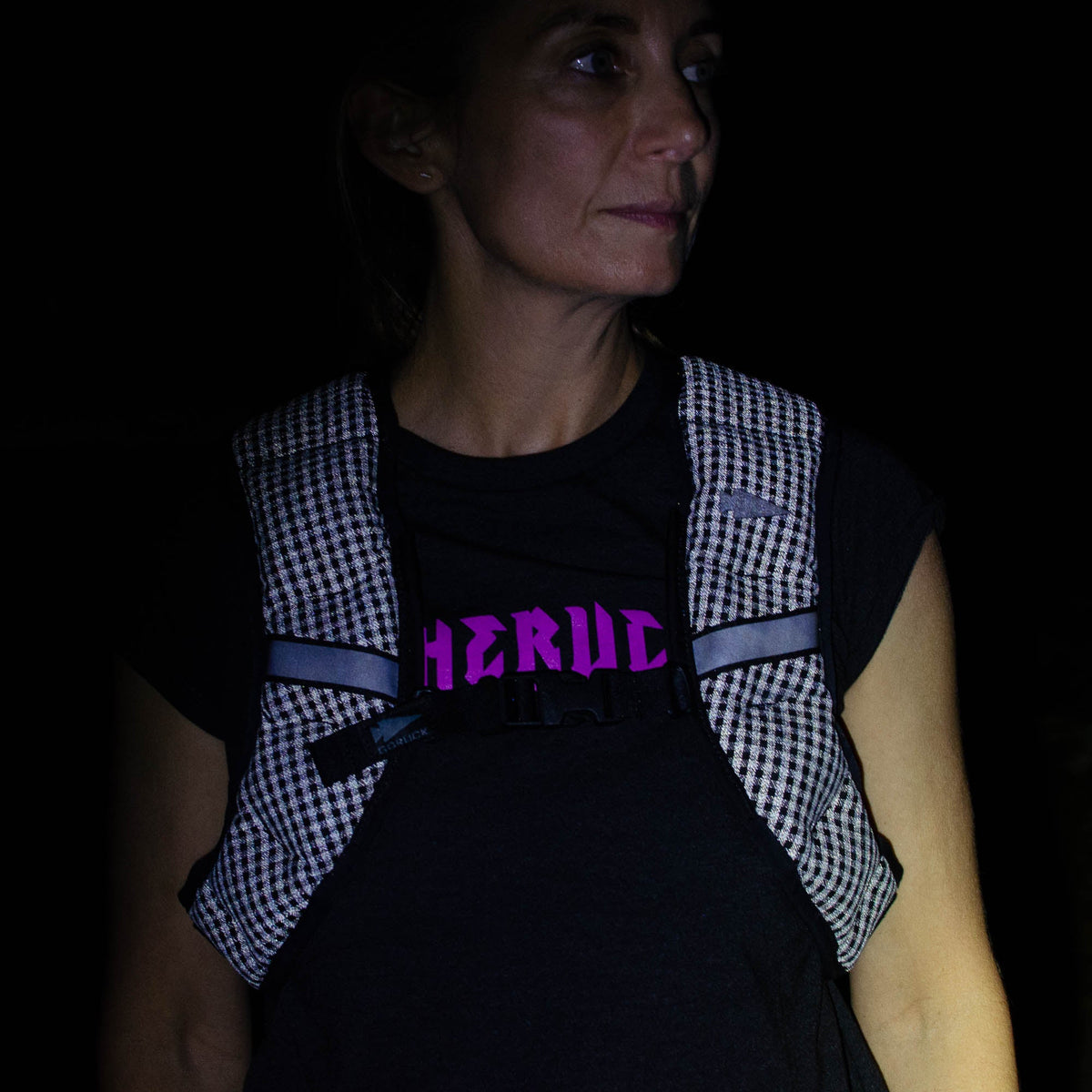 A woman wearing the Spy Ruck | Women's Weighted Vest over a black shirt with "HERCUL" in pink text stands in the dark.