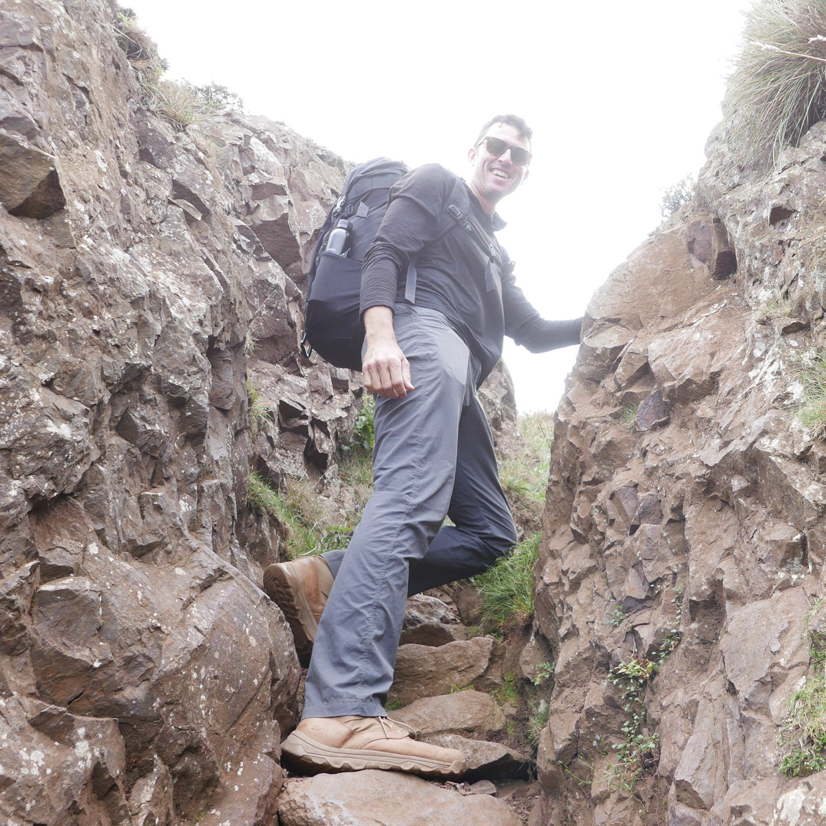A man with a backpack hikes between rocky cliffs, smiling at the camera on a cloudy day, wearing rugged MACV-2 - Mid Top - Briar + Coyote boots designed for adventure.
