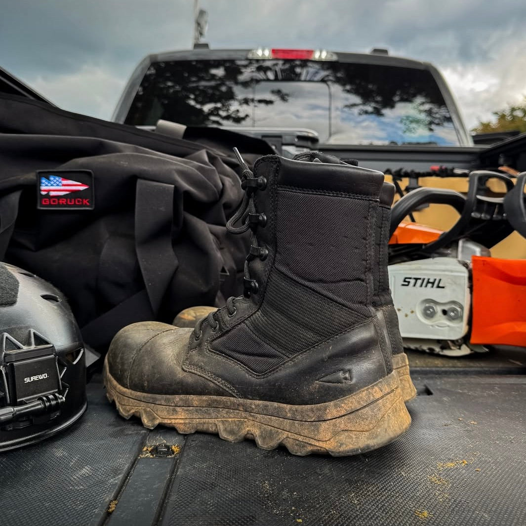 The MACV-2 Safety Boot - 8" High Top sits muddy and black on a truck bed, surrounded by work gear, a helmet, and a Stihl chainsaw in the background.