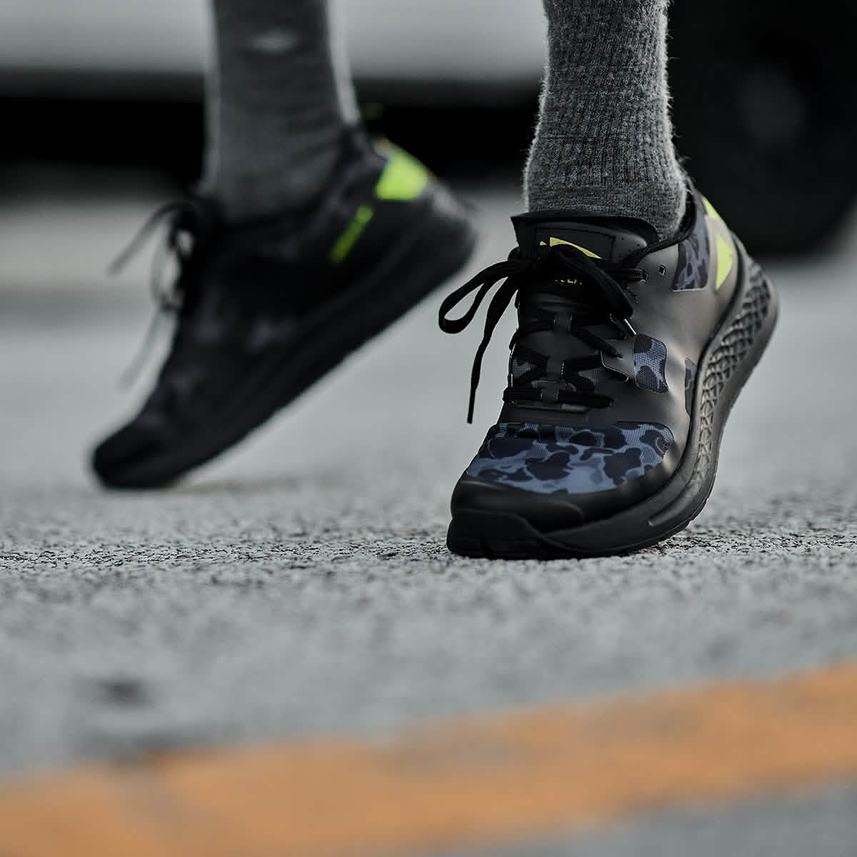 Close-up of a person wearing the Rough Runner - Midnight Frogskin + Acid Lime by GORUCK, showcasing black and green athletic shoes with a camouflage pattern and EVA Midsole, running on a textured outdoor surface. The focus is on the shoes, with legs in gray socks visible.