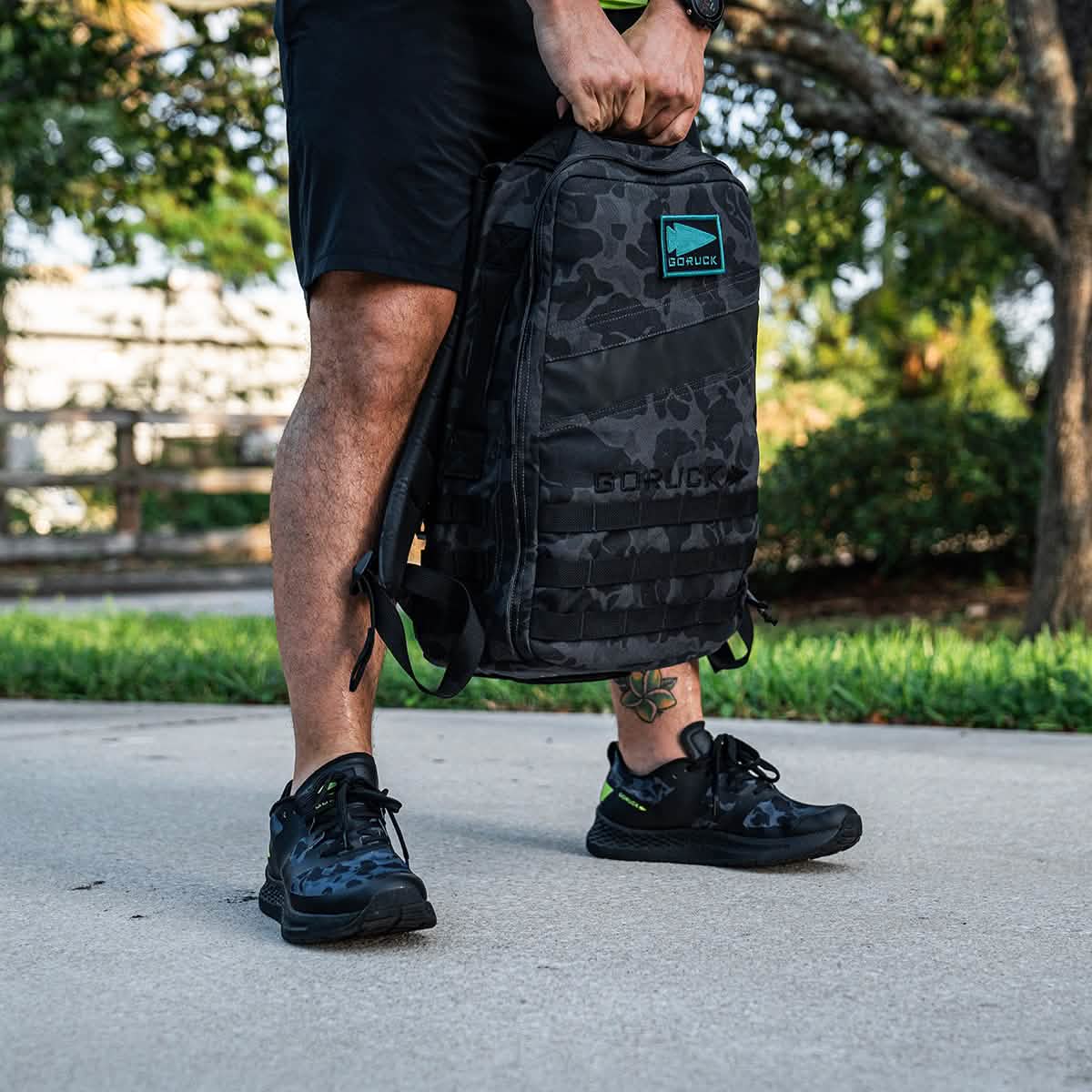 A person in black shorts and sneakers, equipped with a GORUCK Rough Runner - Midnight Frogskin + Acid Lime backpack, stands on a paved path. The scene features green grass and a tree in the background, blending the rugged spirit of adventure with serene nature.