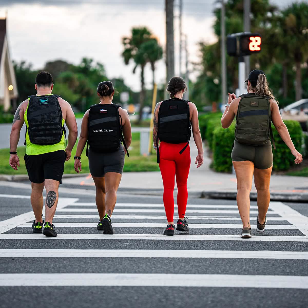 Four individuals equipped with Rough Runner - Midnight Frogskin + Acid Lime backpacks from GORUCK make their way across the street at a crosswalk. The illuminated walk signal guides them under the sun, as palm trees sway alongside contemporary architecture, embodying an active spirit and hinting at the energetic essence of Rough Runner® in this dynamic outdoor scene.