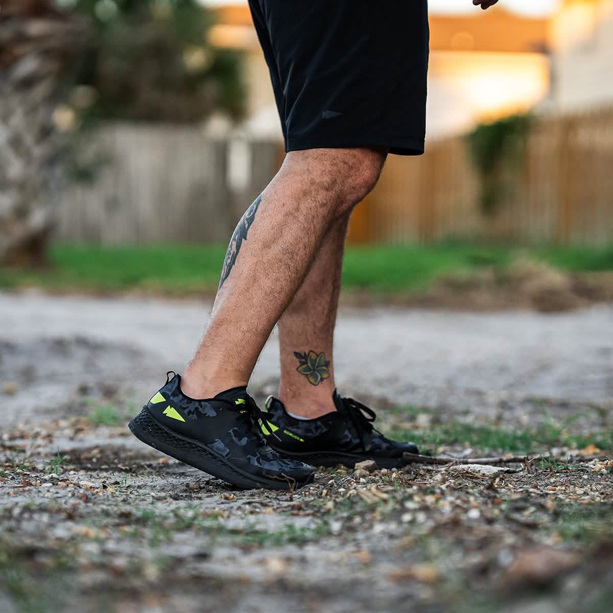 A person donning black shorts and GORUCK's Rough Runner - Midnight Frogskin + Acid Lime sneakers strides along a dirt path. Their tattooed legs add character to the scene, framed by a wooden fence and out-of-focus greenery, creating the perfect backdrop for this energetic walk.