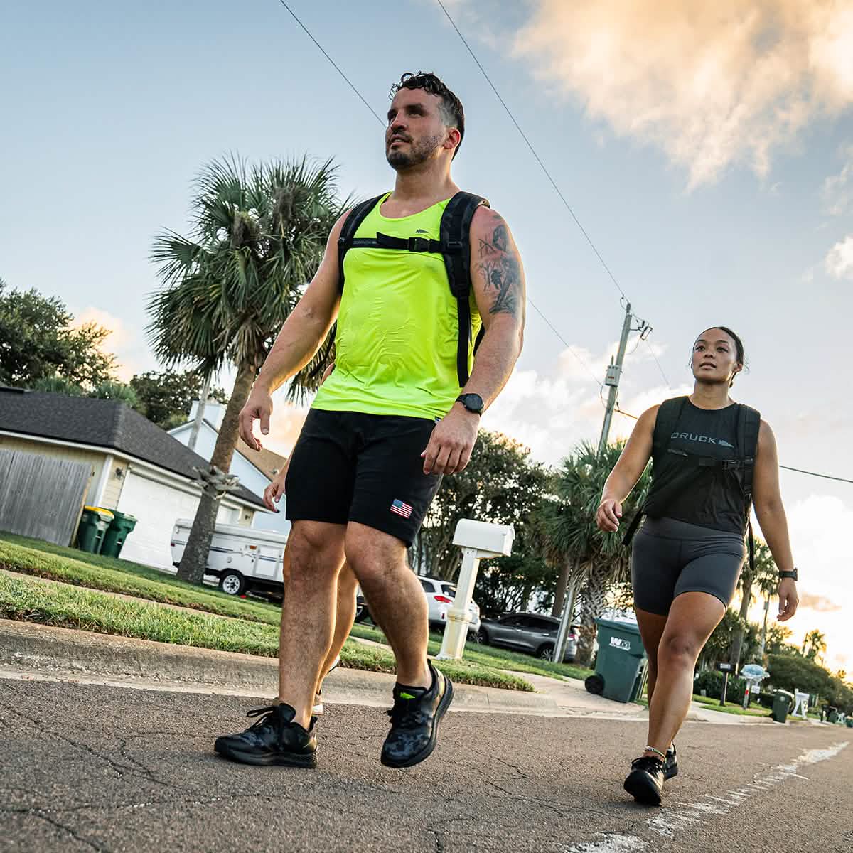 As the sun sets, two individuals walk briskly down a suburban street, outfitted in athletic gear and carrying backpacks from Rough Runner®. The man is dressed in a neon tank top and shorts from GORUCK's Rough Runner collection in Midnight Frogskin + Acid Lime, while the woman opts for a black tank top and shorts. The scene is framed by palm trees and houses, creating a picturesque view.