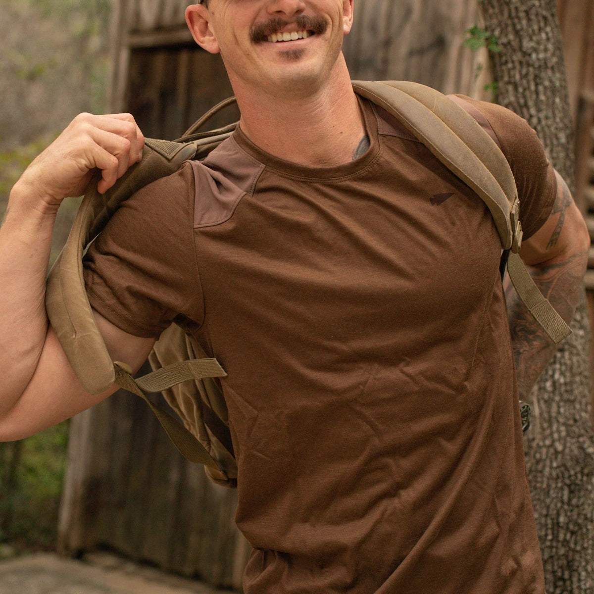 Man wearing GORUCK brown rucking shirt and backpack outdoors near wooden building