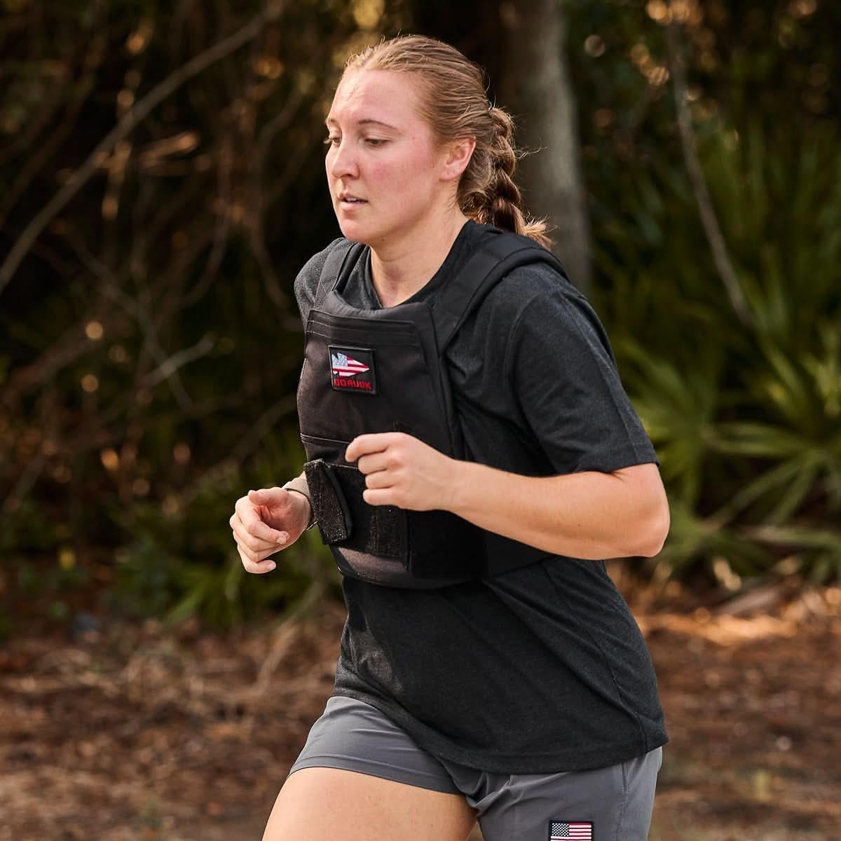A woman runs outdoors in athletic gear and a Rucking Weight Vest with Curved Ruck Plates, surrounded by lush greenery—perfect for rucking enthusiasts.