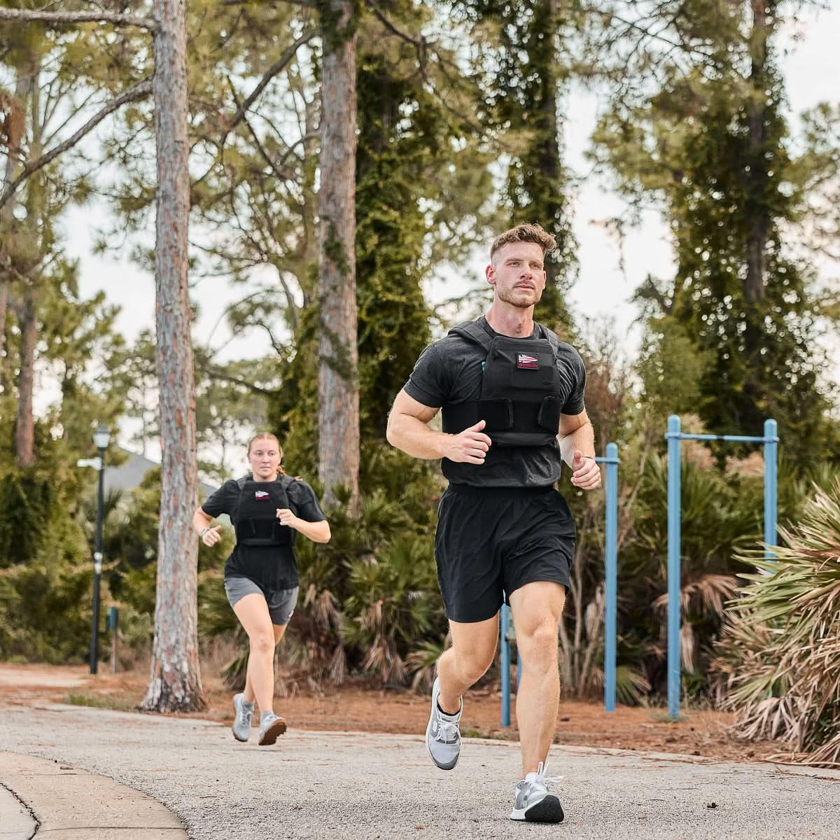 Two people running outdoors wearing GORUCK training vests on a forest trail