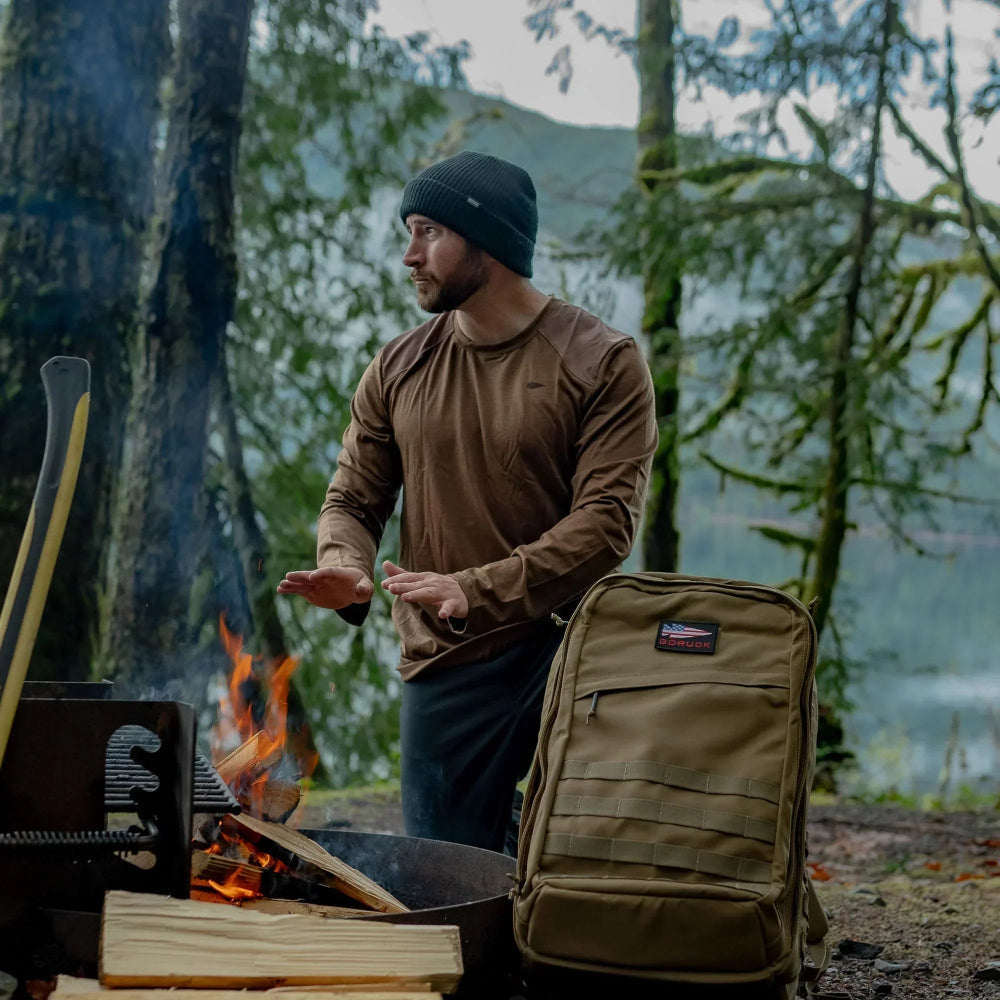 A man wearing the Men’s Commando Long Sleeve - Merino Wool shirt warms his hands by a forest campfire, with a backpack and chopped wood beside him.