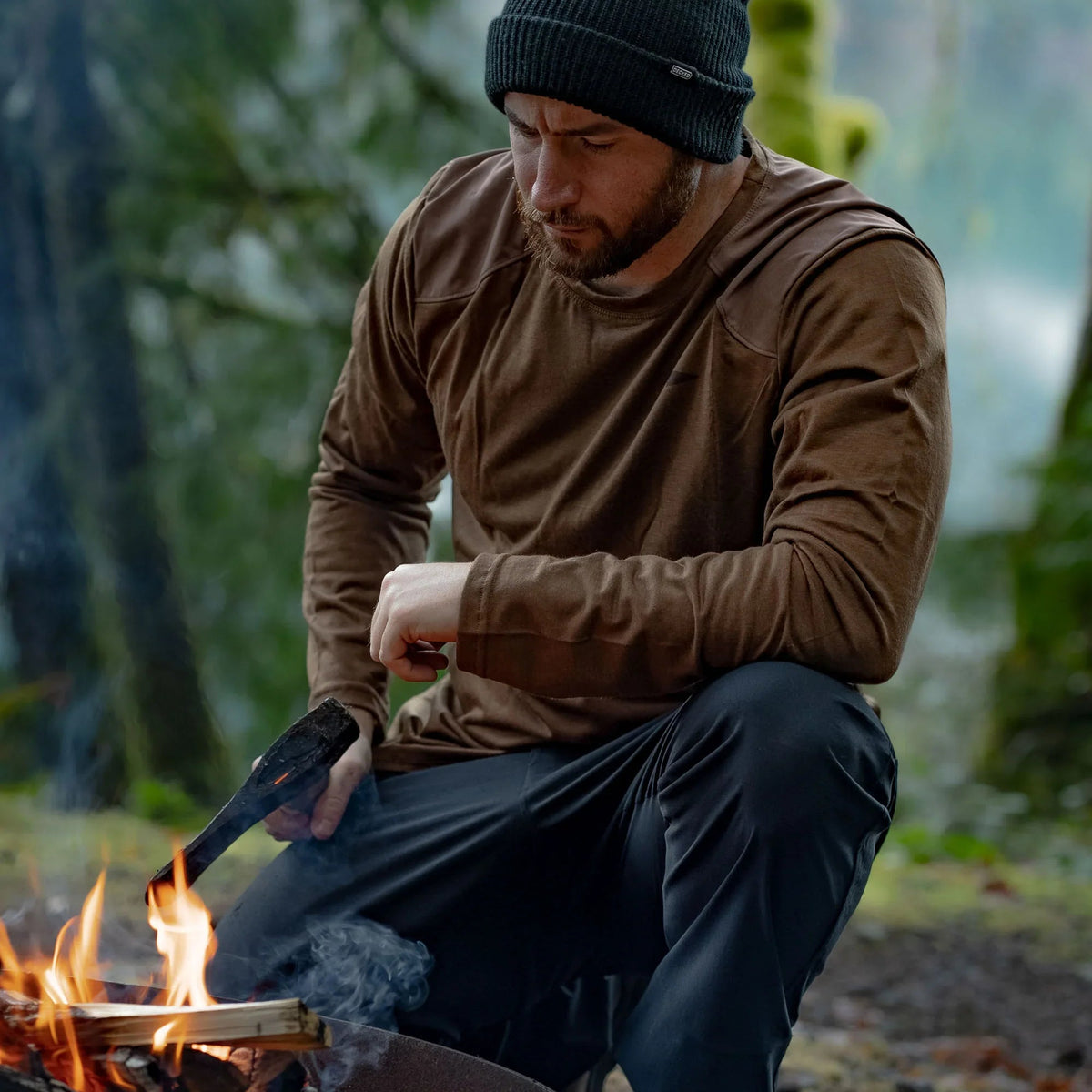 Man in GORUCK outdoor gear tending campfire in forest, wearing brown shirt and black beanie.
