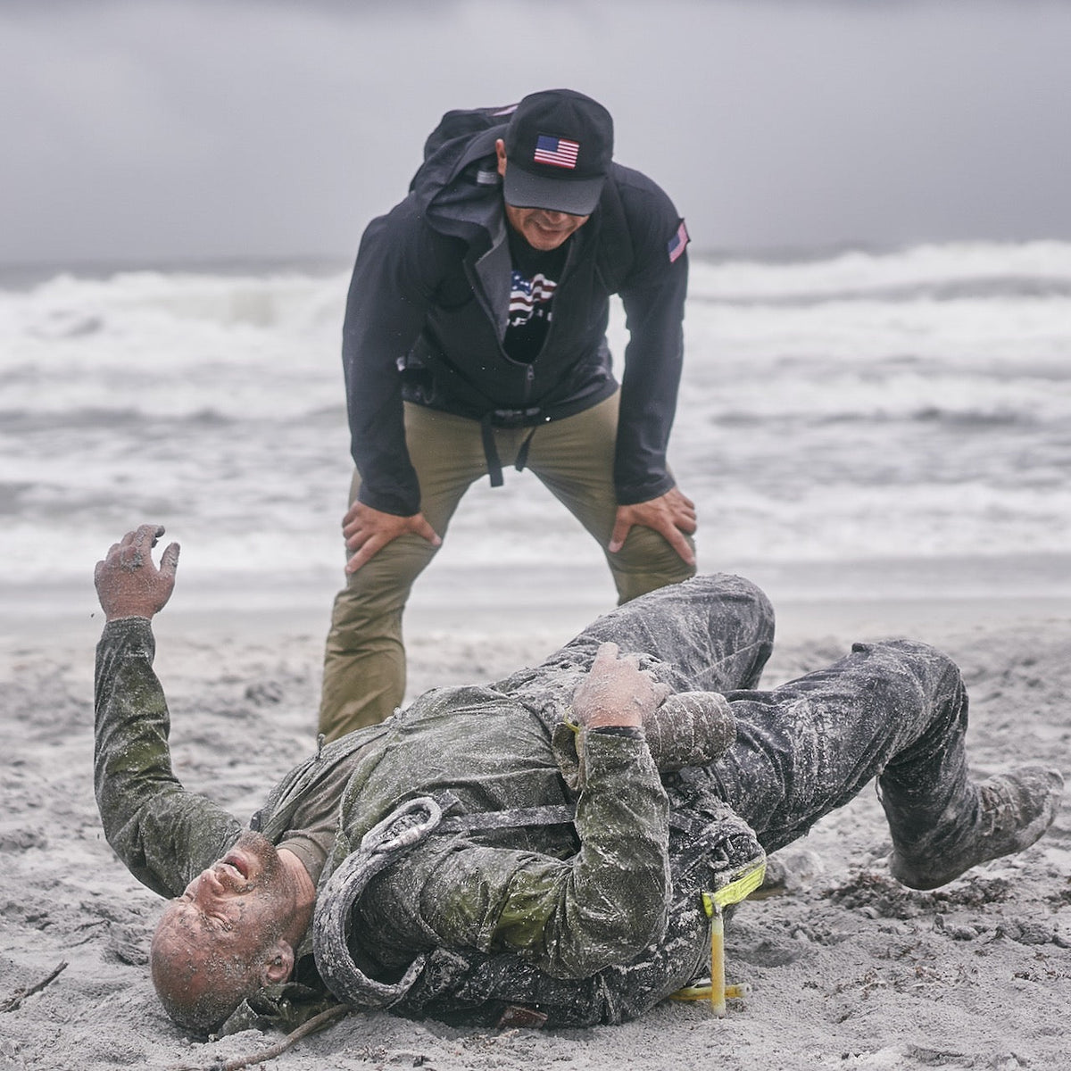 A man in an American flag cap and abrasion resistant pants watches over another man covered in sand lying flat on his back on a beach.