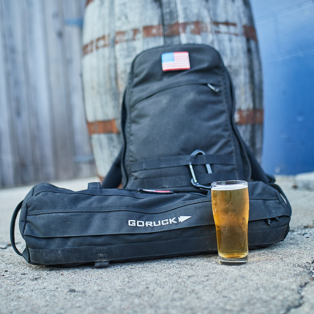 A backpack and GORUCK bag with a glass of beer sit on the ground in front of a barrel, next to a Sandbag Kit (w/ Filler Bag) built for serious training.
