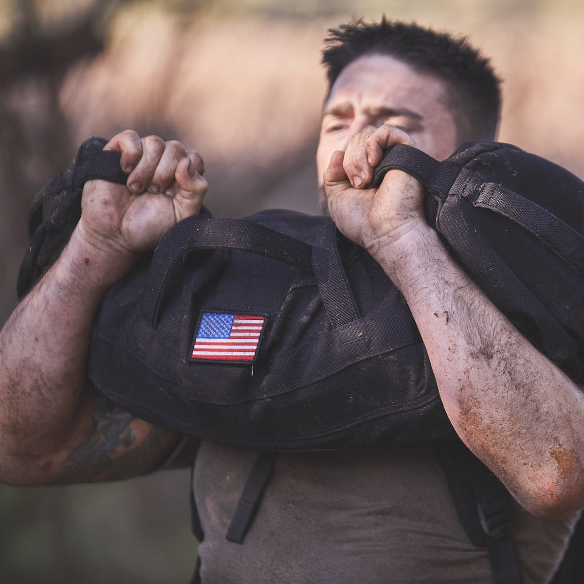 A person with muddy arms lifts a Sandbag Kit (w/ Filler Bag) featuring an American flag patch outdoors.