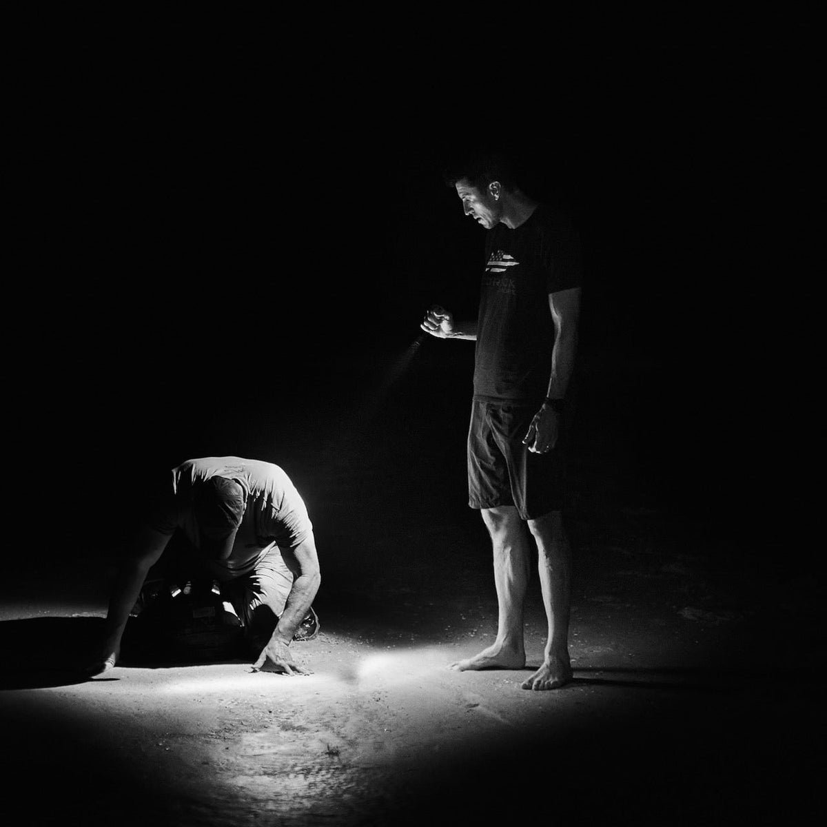 Two men in the dark; one stands with a flashlight, the other kneels in its beam, wearing Men’s Simple Shorts - Lightweight ToughDry® made from 2-way stretch fabric for comfort in challenging conditions.
