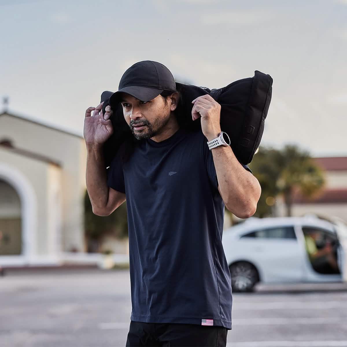 Man wearing GORUCK black hat and shirt lifting rucking sandbag outdoors in parking lot