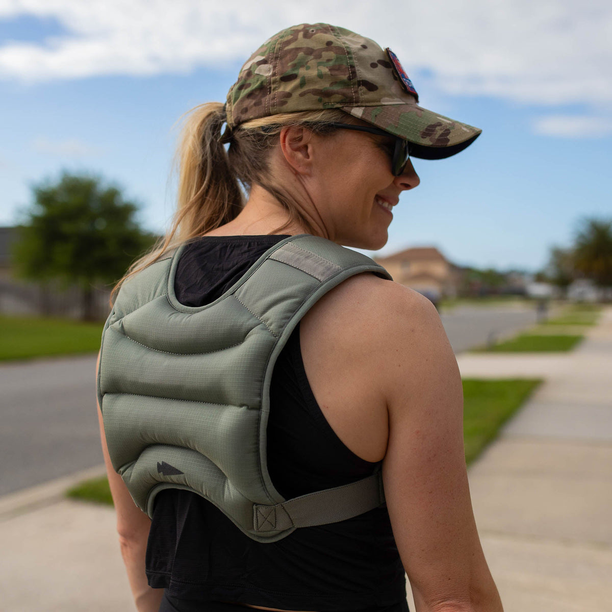 A woman wearing a camouflage hat and the Spy Ruck | Women's Weighted Vest walks outside on a sunny day.