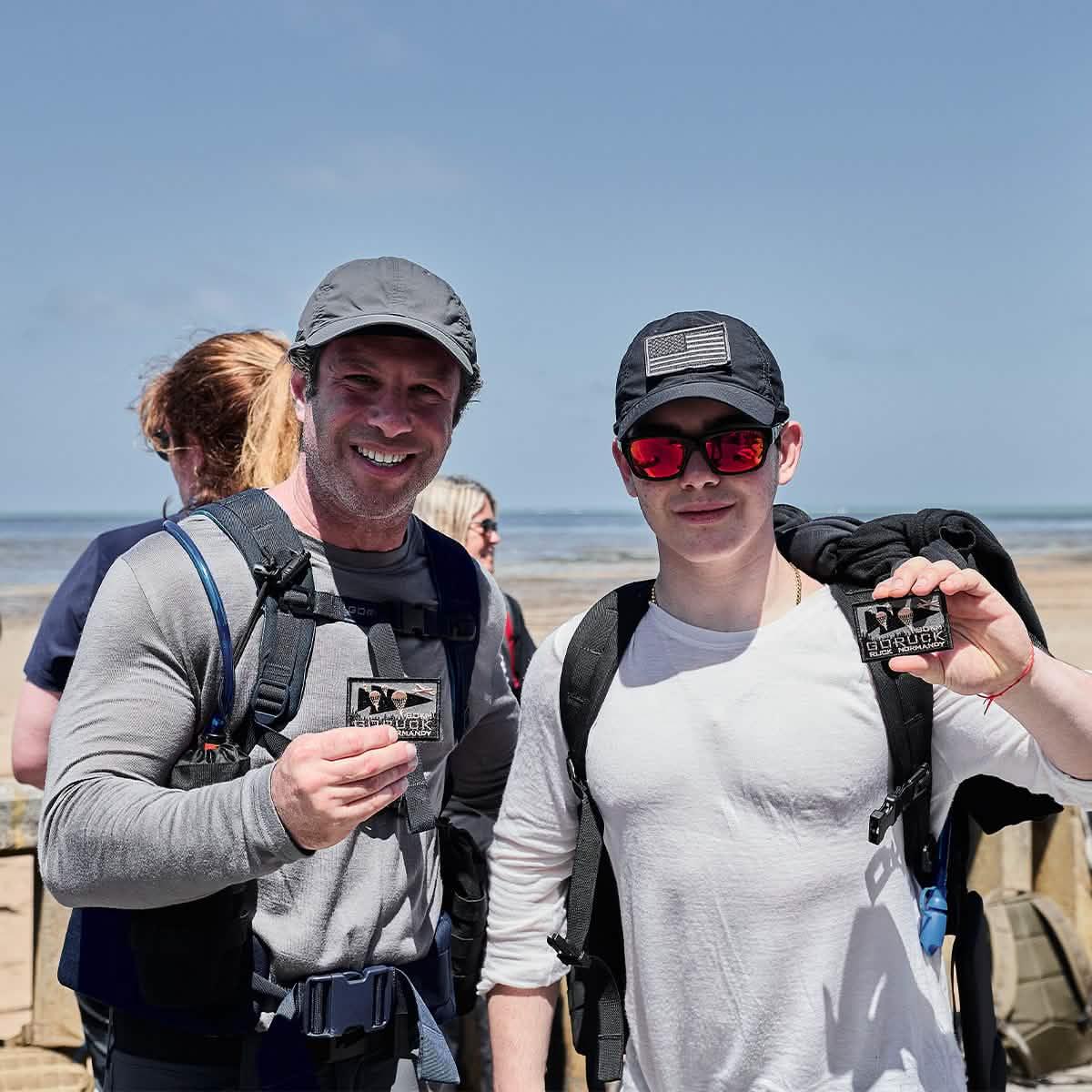 Two men in GORUCK gear with backpacks and patches on a sunny beach, smiling after rucking event