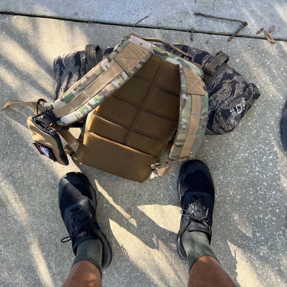 A person wearing Mackall - Black + Forged Iron sneakers stands near a tactical backpack and a camouflage bag on concrete.
