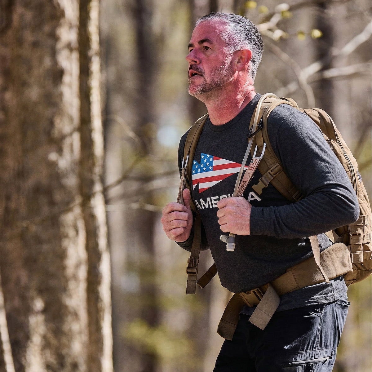 A man hikes in the woods wearing the Men's America Long Sleeve - Tri-BlendX and a backpack with an American flag design, looking focused.