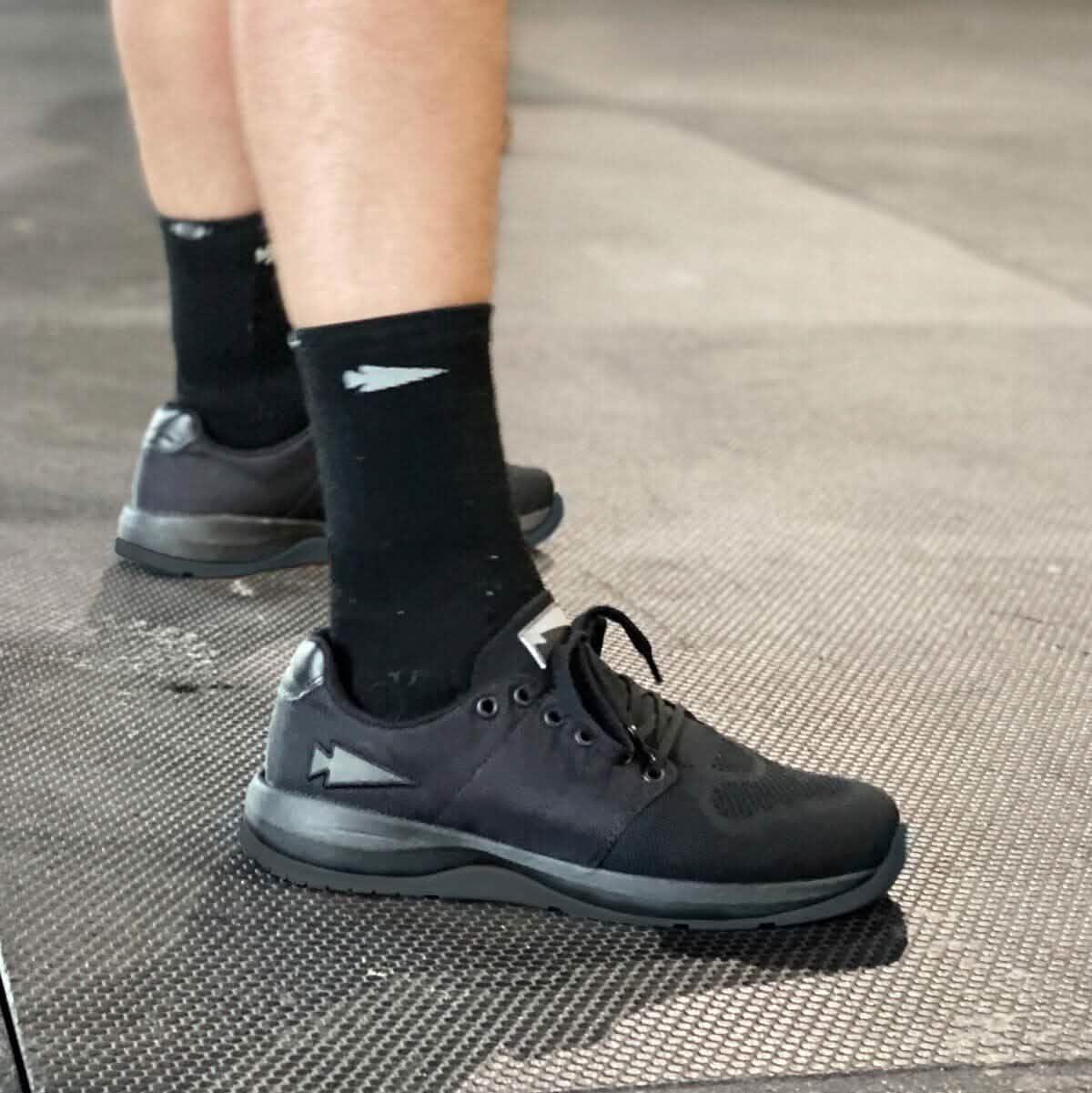 A person stands on a textured floor displaying alternating light and dark sections, ideal for showcasing functional fitness footwear. They are wearing GORUCKs Ballistic Trainers - Blackout, made with CORDURA® Ballistic Nylon and 3X Support™, paired with black socks featuring a white arrow design.