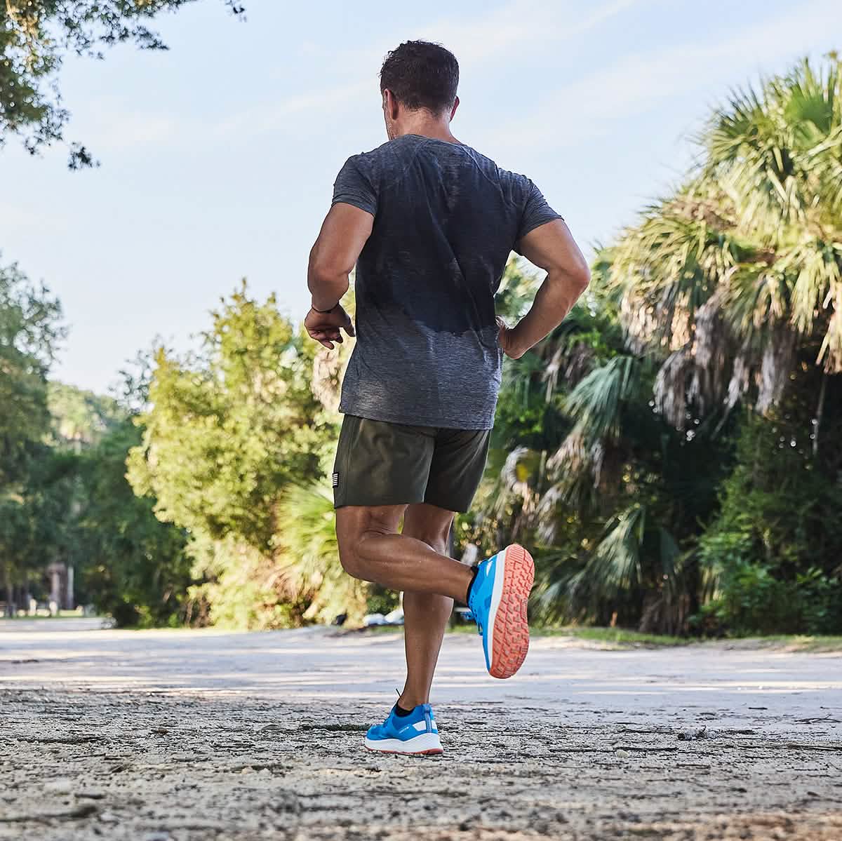 A man is jogging along a sunlit path enveloped by lush greenery, exemplifying the essence of fitness and nature. Wearing GORUCK's Rough Runner - Electric Blue shoes with orange soles, paired with a dark gray shirt and olive shorts, he truly embodies the spirit of an outdoor enthusiast.