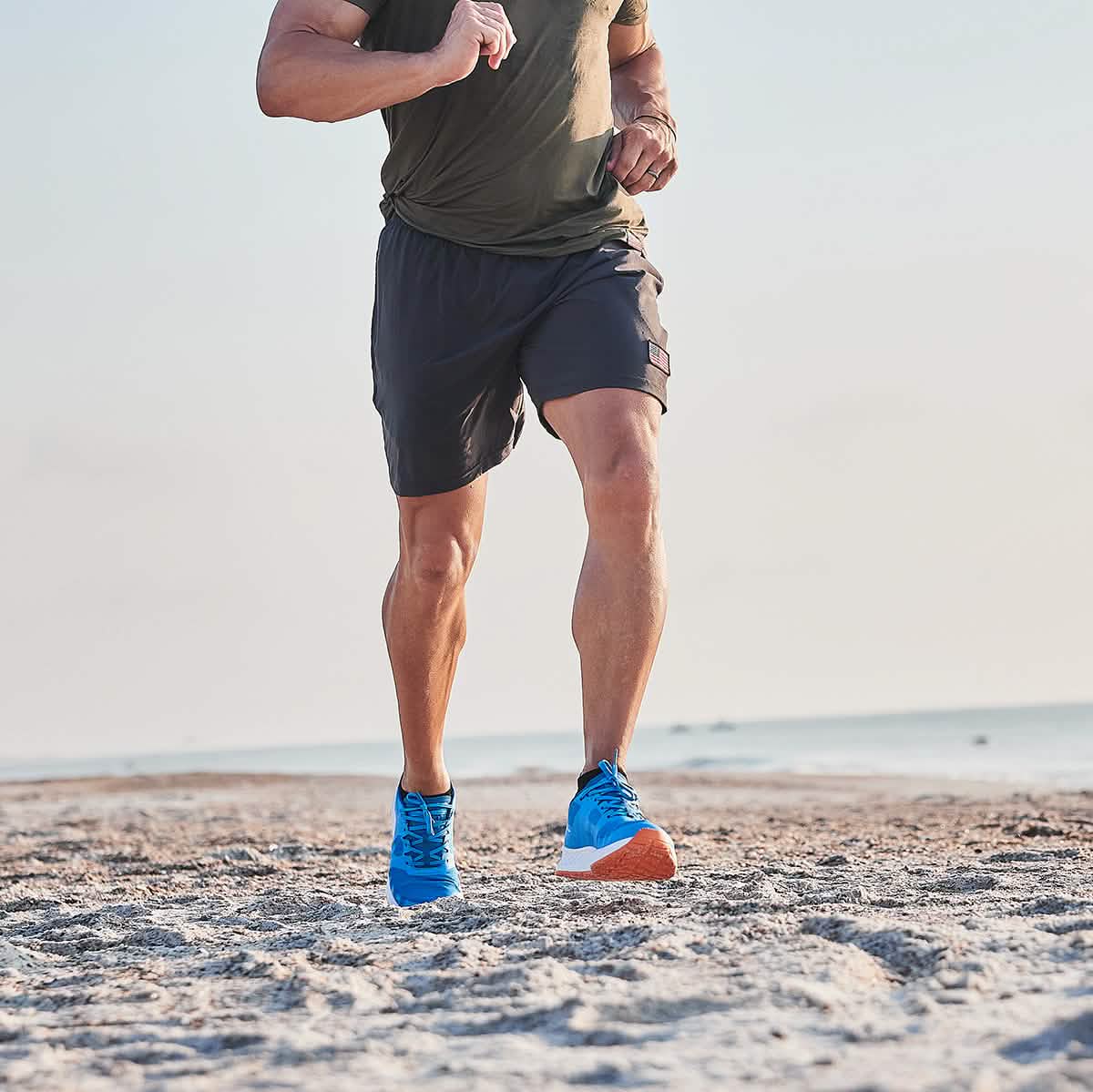 A person jogging on the beach, clad in a green shirt and dark shorts, wearing the Rough Runner - Electric Blue from GORUCK. The Rough Runner enjoys the clear sky as the ocean graces the background with its serene presence.