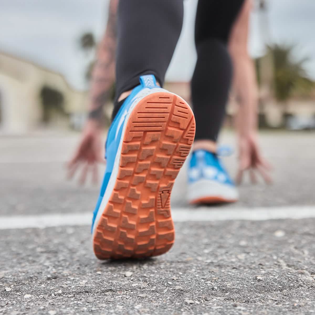 A person in running gear is in a crouched starting position on an outdoor track, highlighting their GORUCK Rough Runner shoes in Electric Blue. These high mileage running shoes feature a textured sole and Gradient Density EVA midsole, engineered for endurance and ideal for the rough runner's journey.