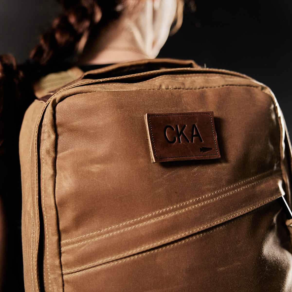 A person with braided hair is seen from behind wearing a brown backpack, which showcases a vendor-unknown Patch - Custom Leather Monogram featuring the embossed initials "CKA." The dark background accentuates the lighter shade of the backpack.