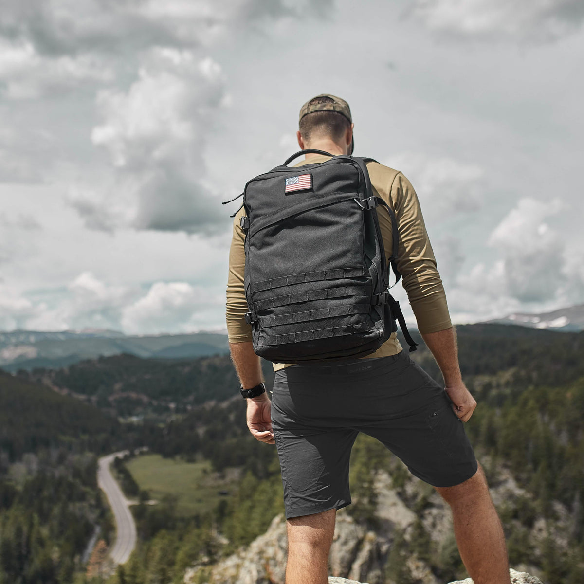 Man wearing a GORUCK backpack with American flag patch, overlooking a scenic mountain landscape.