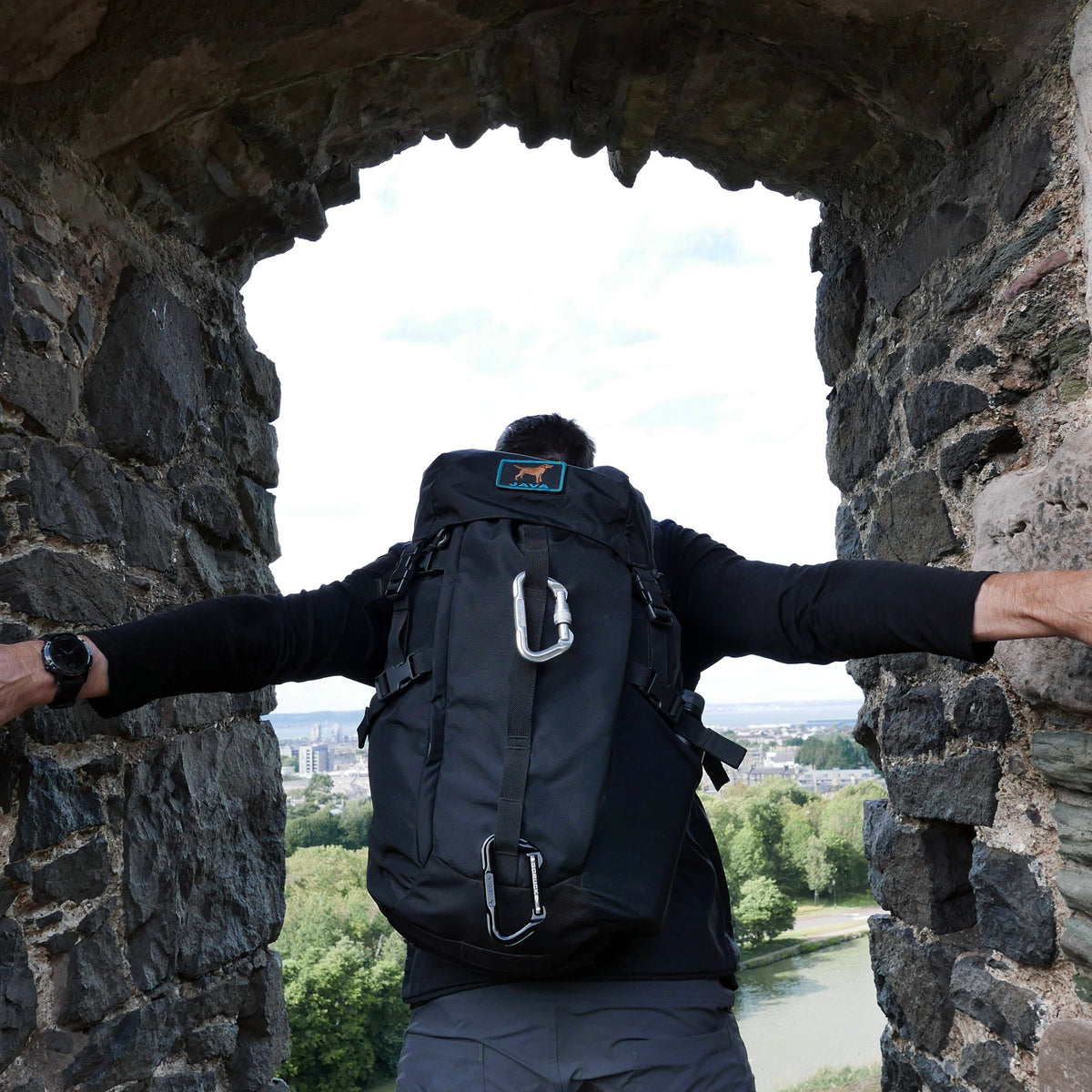 A person with a large black GR MONSTER Rucksack - PREORDER stands in a stone arch, overlooking trees and a distant city.