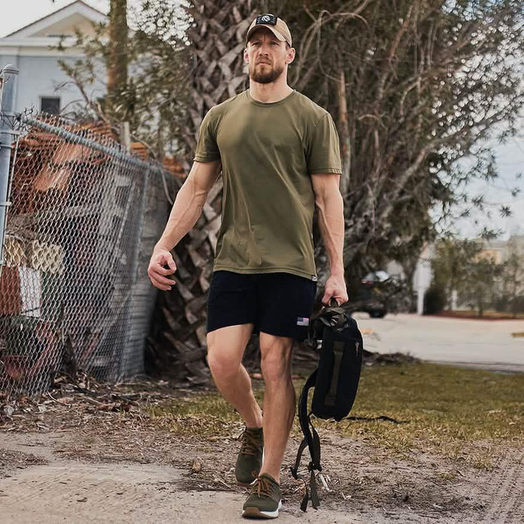 Man in GORUCK shirt and shorts walking outdoors with a rucksack, showcasing rugged rucking gear