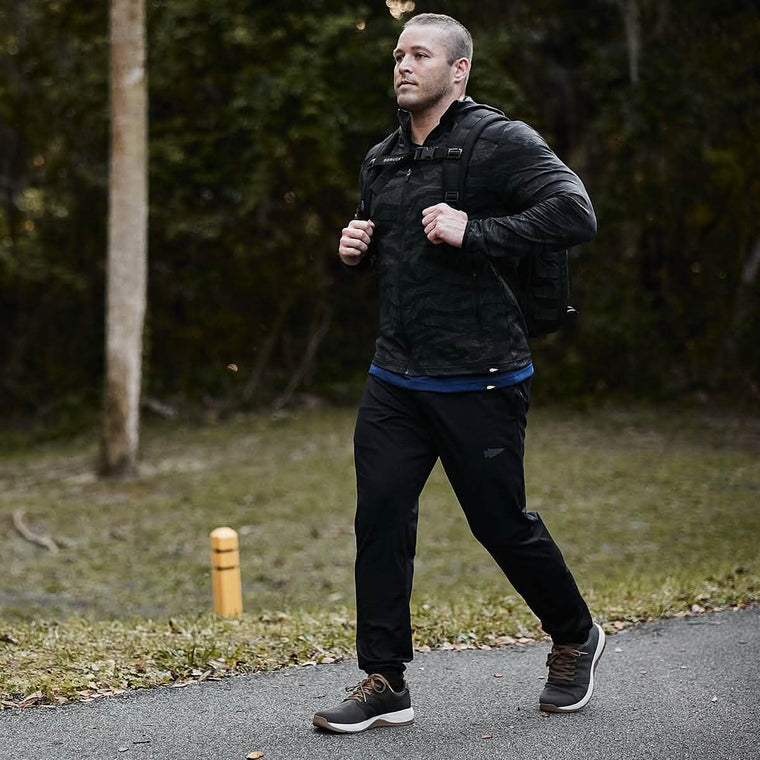 A man walks along a path in a wooded area, wearing a dark jacket and GORUCK's Indestructible Performance Joggers with sneakers. His backpack bears the Scars Lifetime Guarantee. The background is textured with trees and a grassy area beside the path.