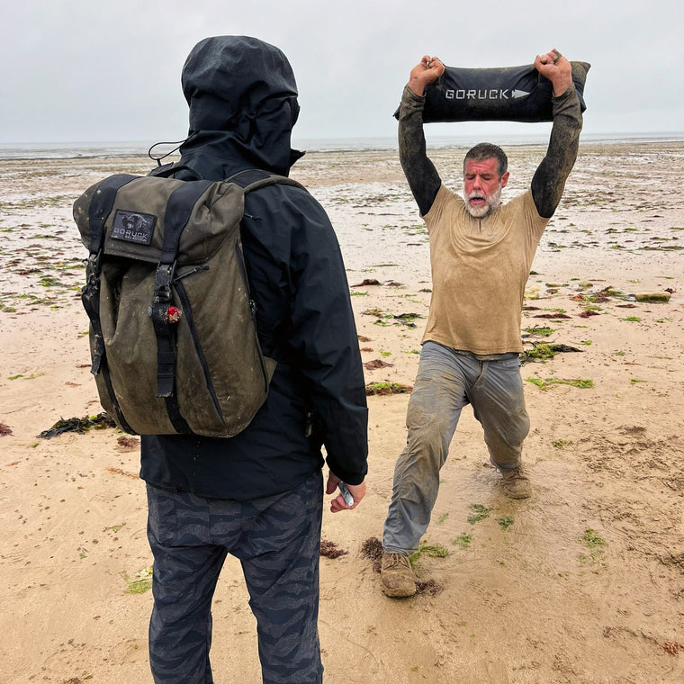 A man in a tan shirt lunges on the beach, lifting a sandbag, while another person with an M26 PREORDER // Q4 2026 ETA pack faces him.